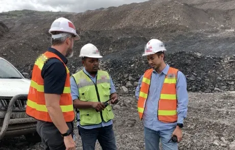Three people in hard hats and hi-vis vests talking beside a haul road at an open-pit mine. Faces are obscured.